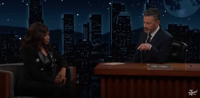 Talk show host interviews a guest on a nighttime set with cityscape backdrop