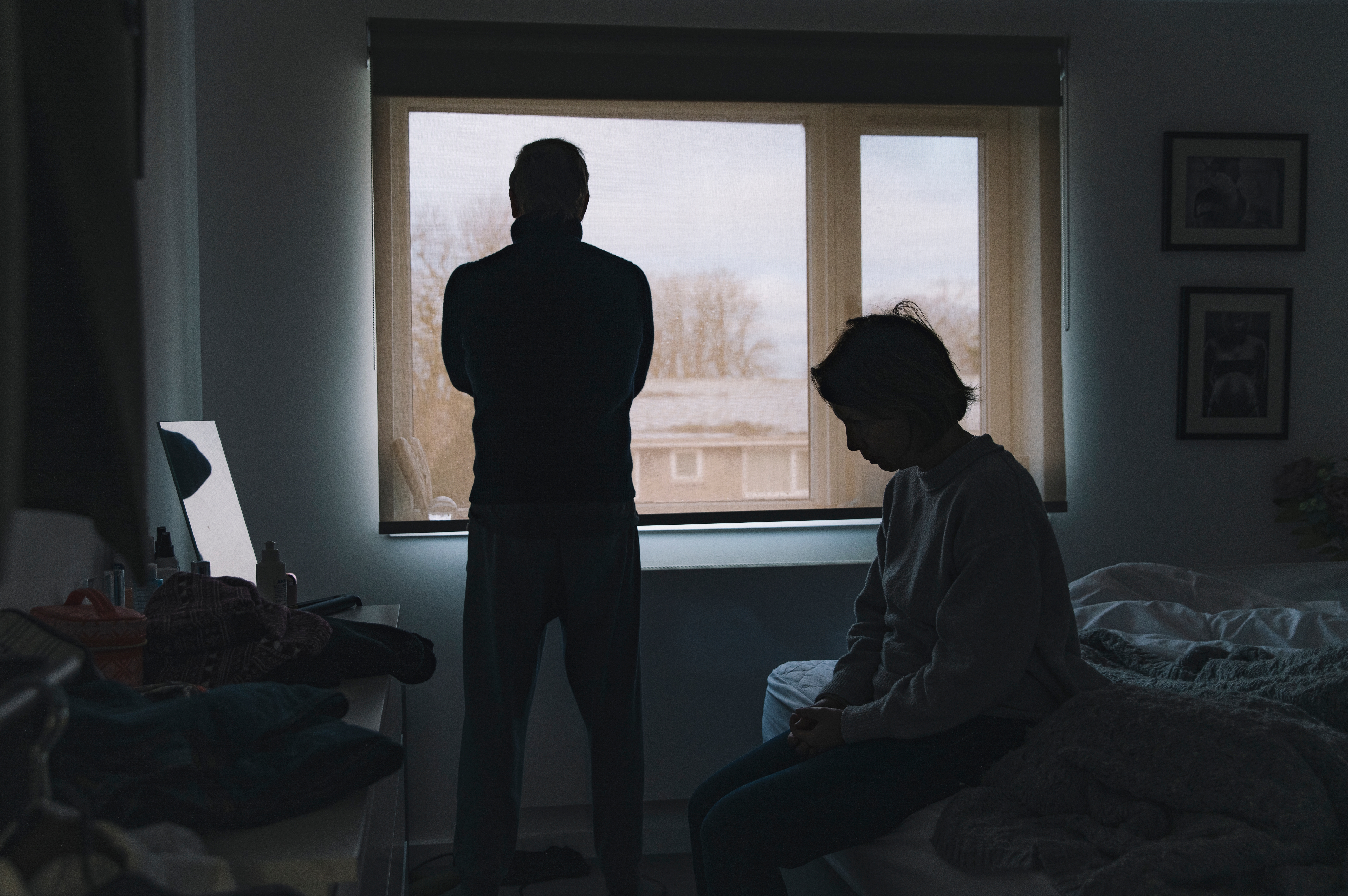 Two people in a dimly lit bedroom, one standing by the window and the other sitting on the bed, both appear to be deep in thought