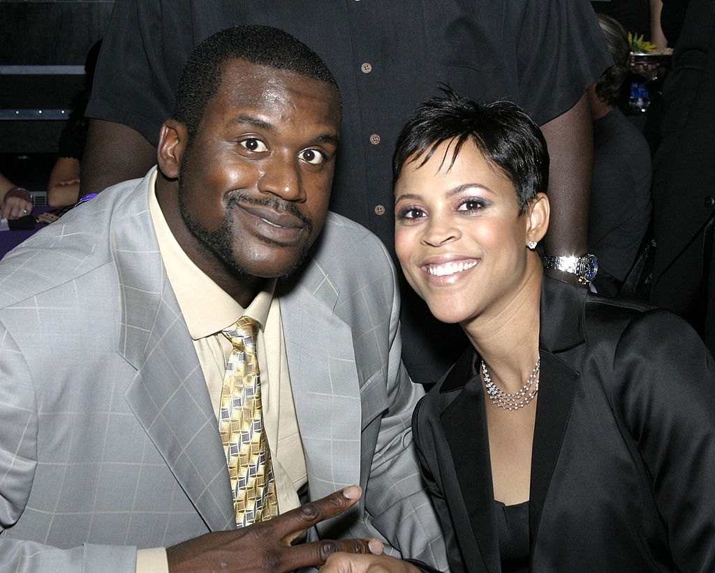 Two people are posing for a photo at an event. One is wearing a suit with a patterned tie, the other is in a sleek black outfit, smiling