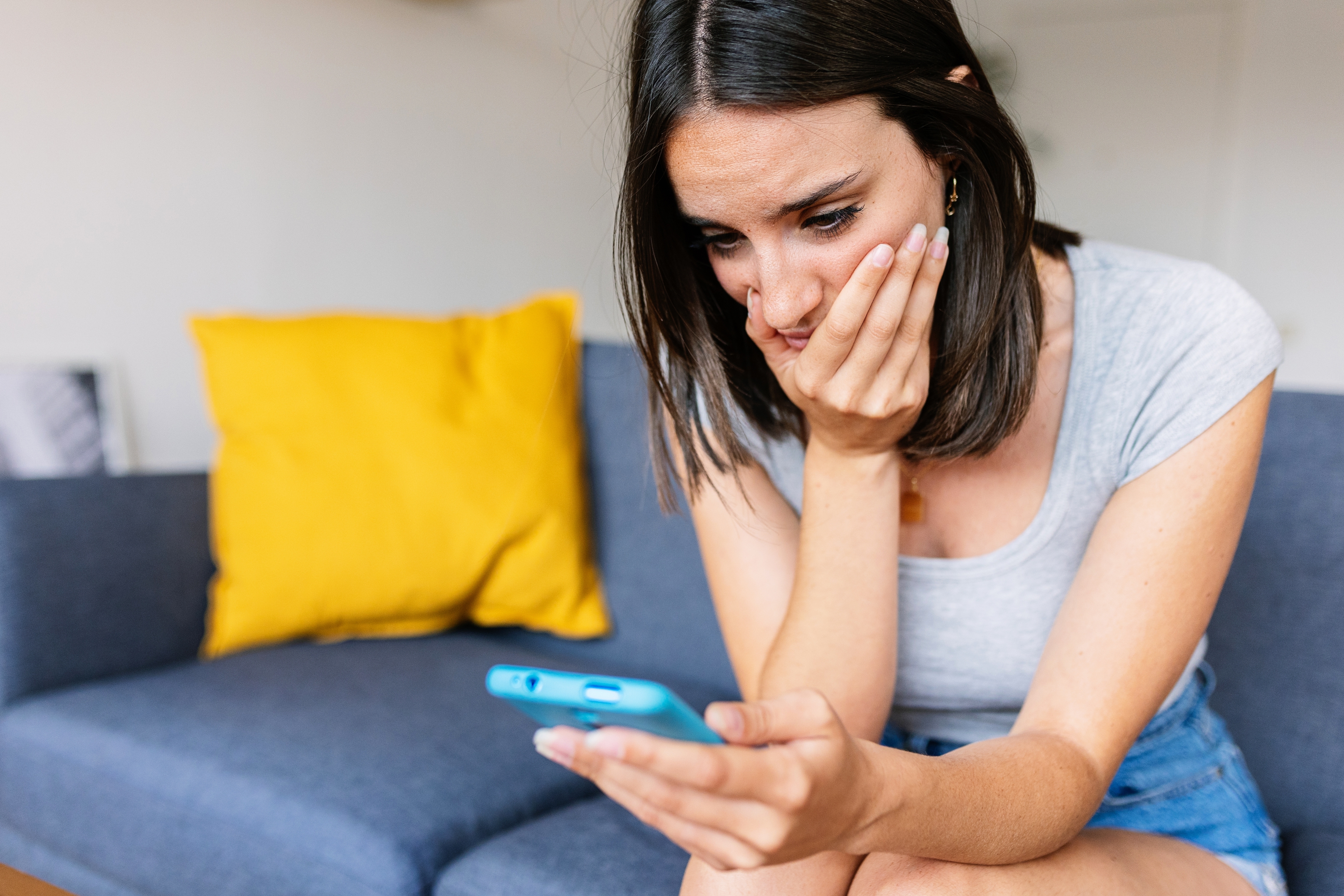 A person sitting on a couch, looking at a phone with a concerned expression, one hand covering their mouth