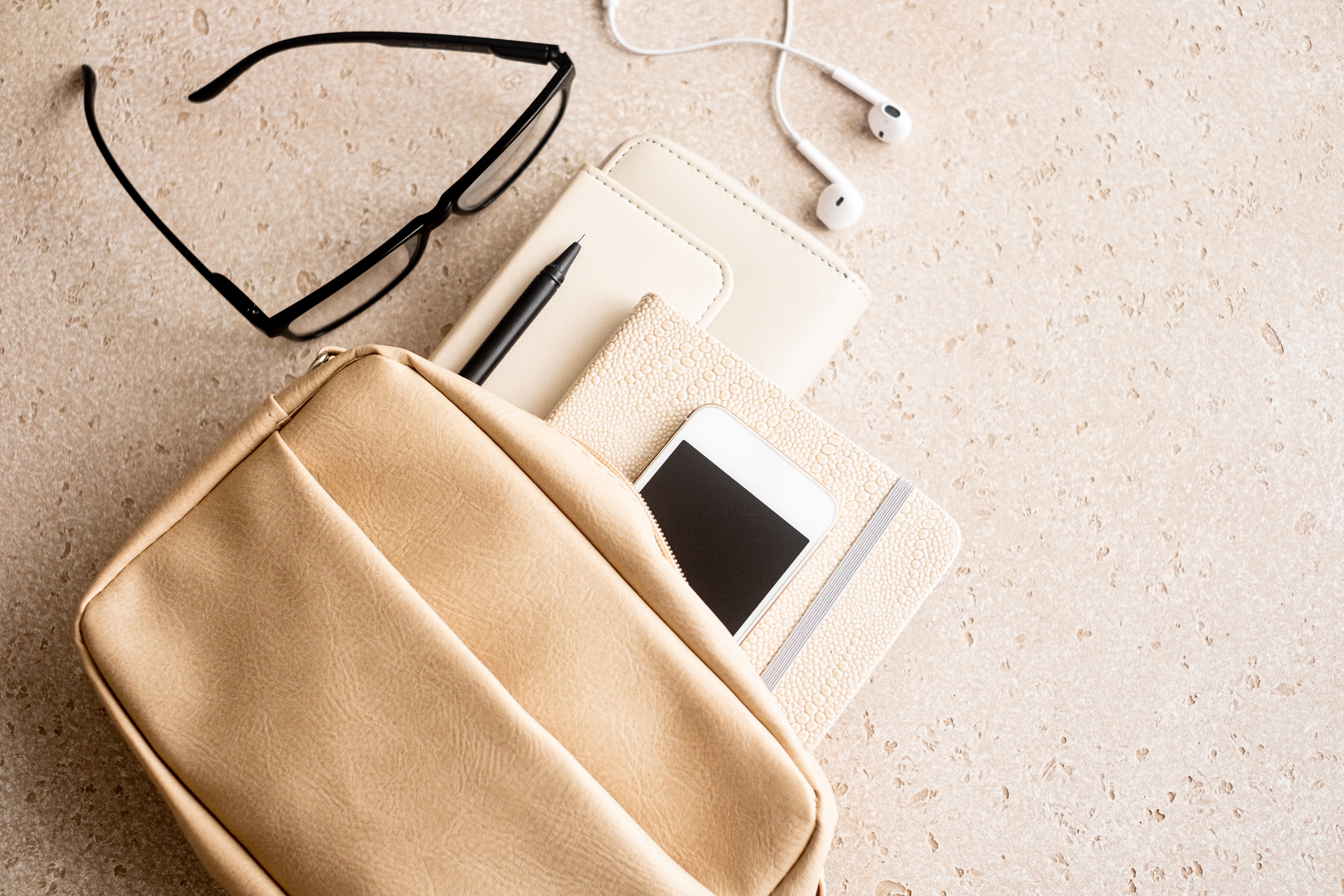 Open bag with glasses, earphones, a pen, a phone, and a notebook on a countertop, suggesting travel or study