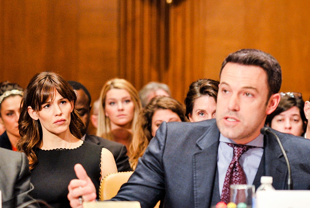 A man in a suit speaks at a hearing; a woman in a sleeveless dress looks on from the background with interest