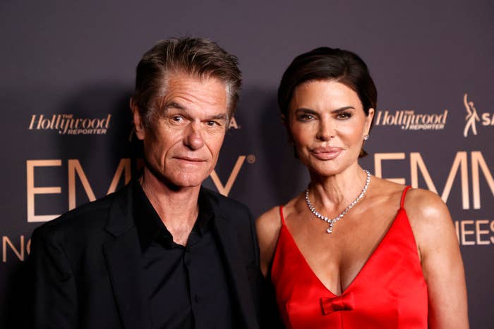 Two people on the red carpet, both in elegant attire. The person on the right is wearing a sleeveless dress and a necklace