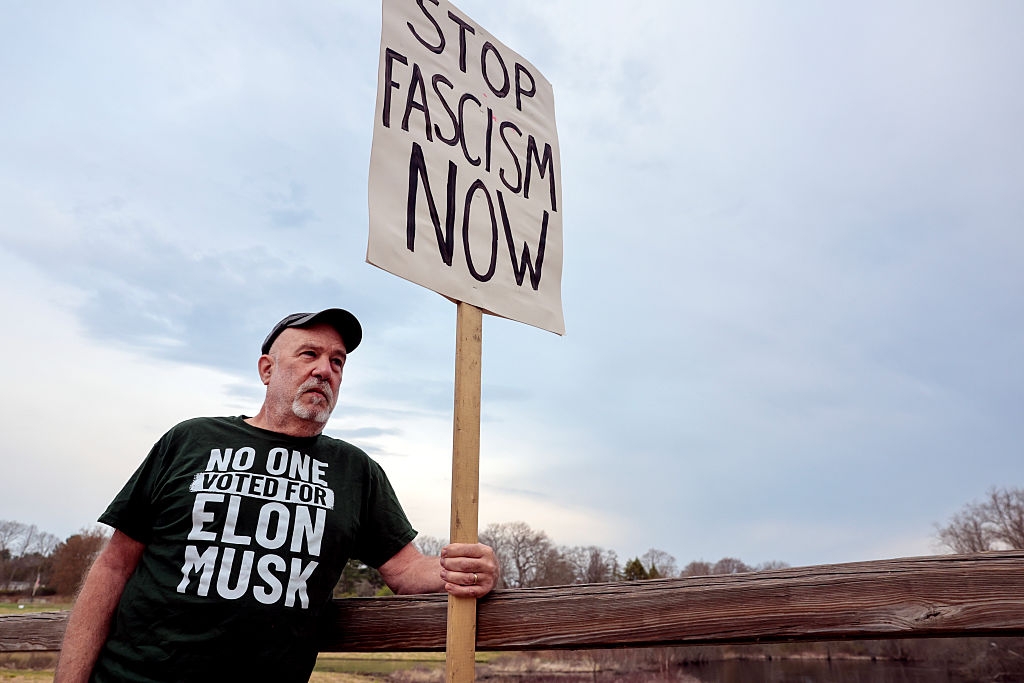A man leans on a wooden fence, holding a "Stop Fascism Now" sign. His shirt reads, "No One Voted for Elon Musk."