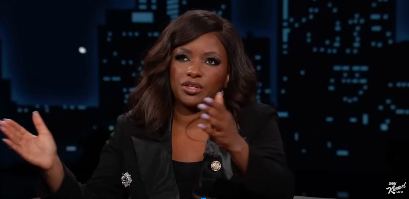 A person appears in a talk show setting, engaged in conversation, wearing a black blazer with a decorative pin