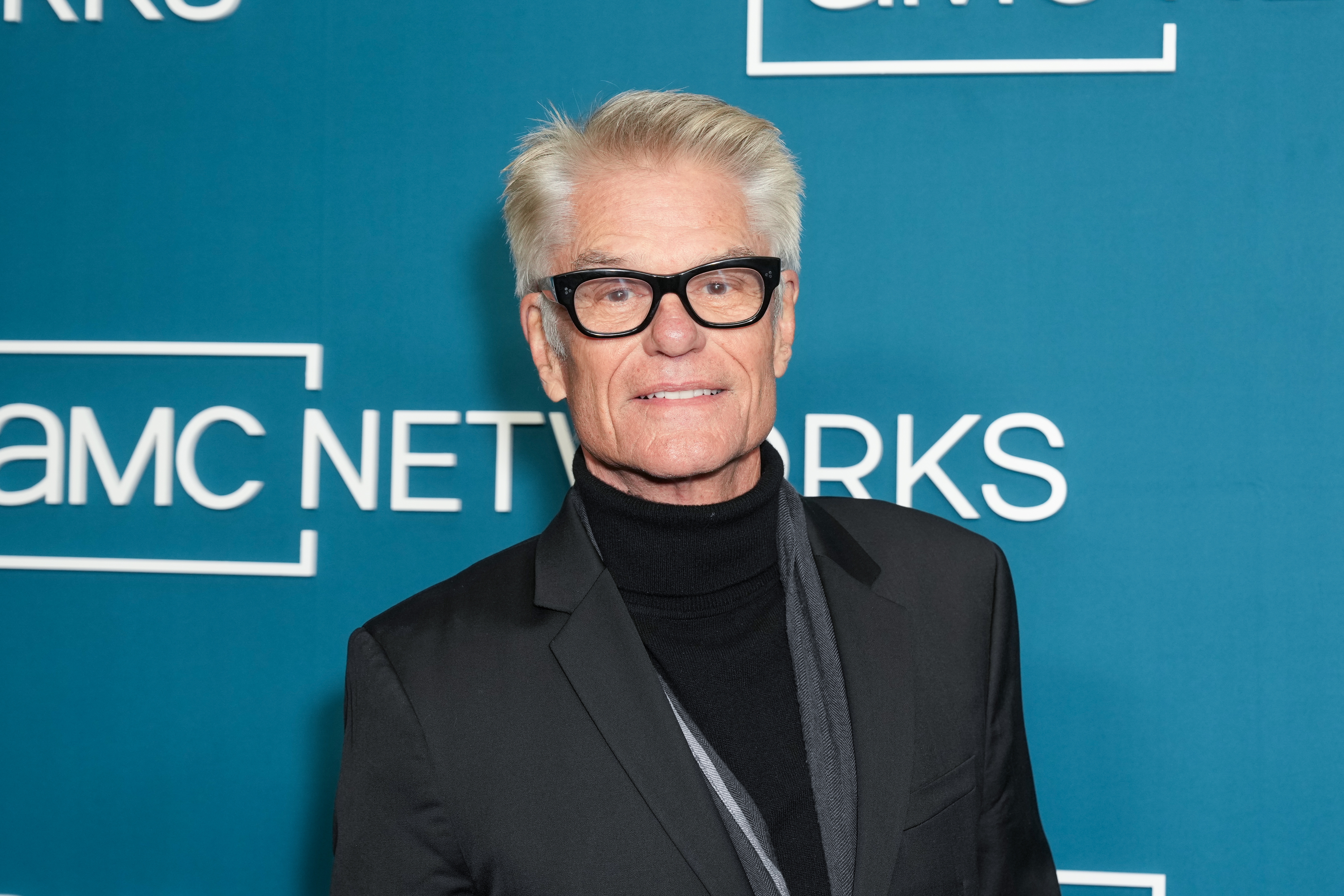 Harry Hamlin in a black suit and glasses on a media event backdrop, smiling