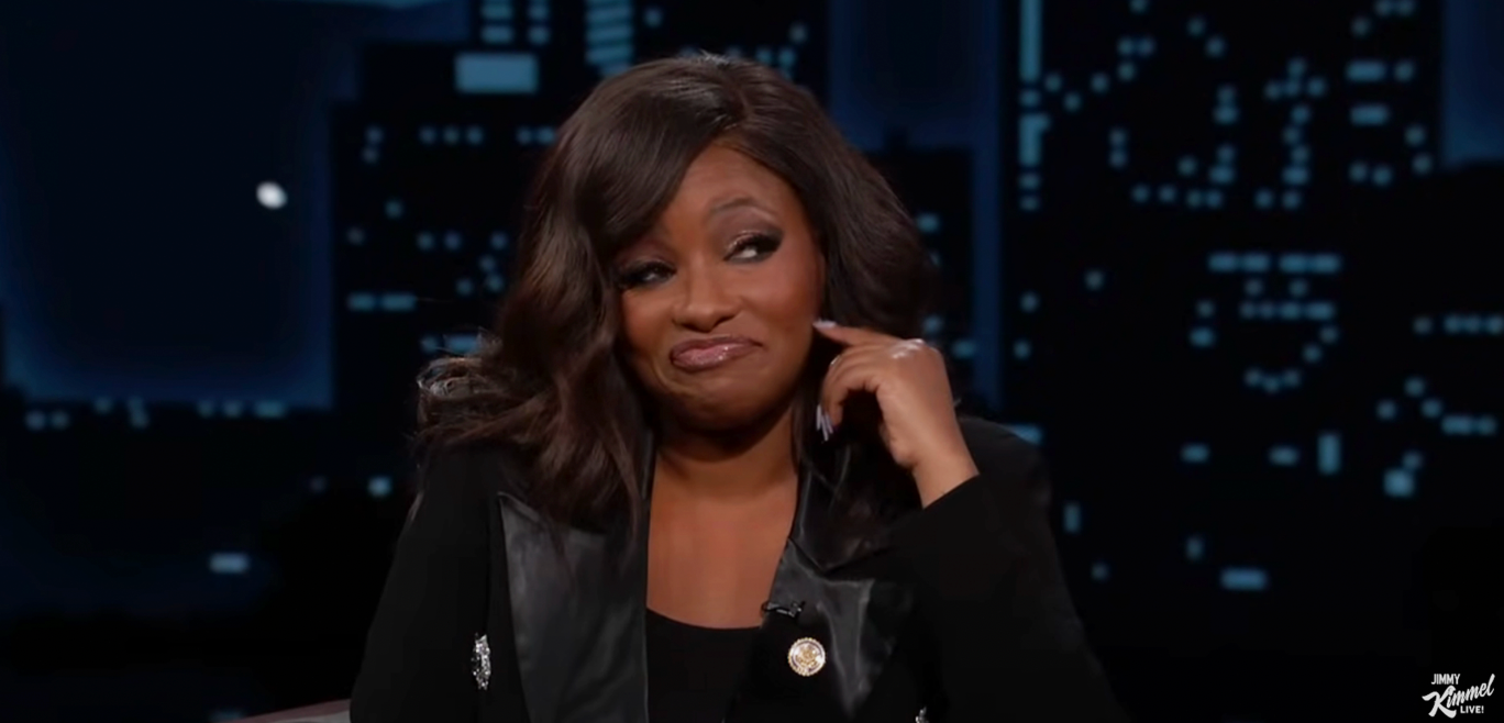 A person on a talk show set, wearing a black jacket, smiles slightly with one eyebrow raised