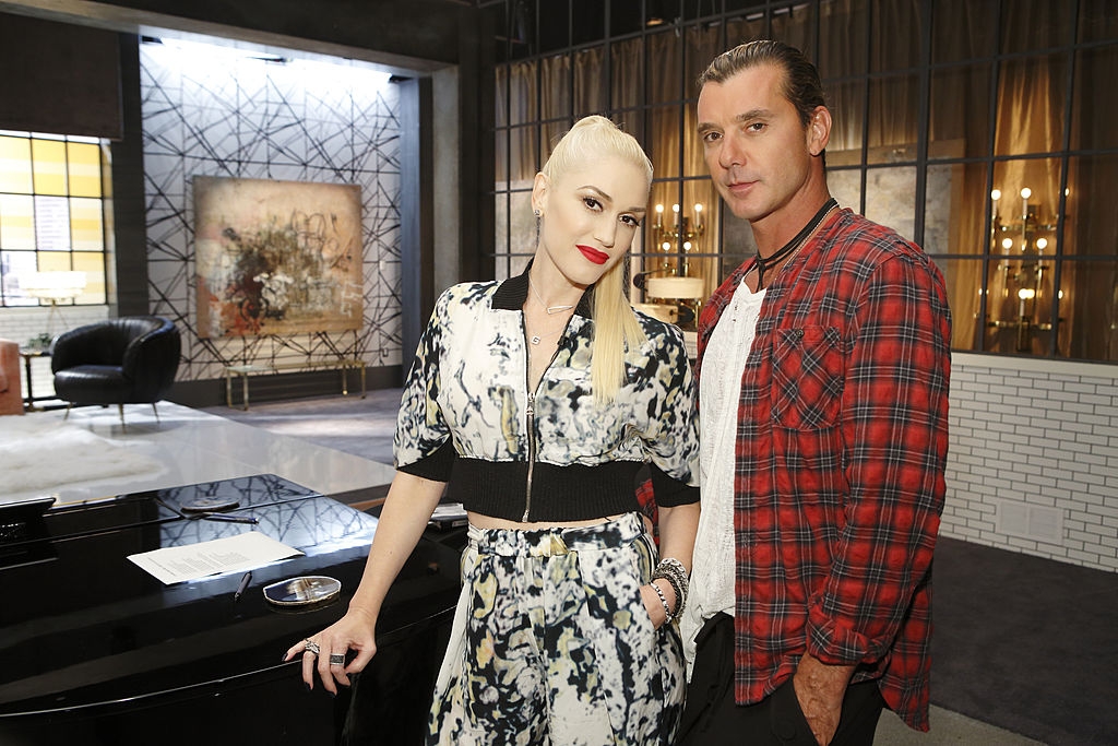 Two celebrities pose together indoors; one wears a patterned jumpsuit, the other a red plaid shirt