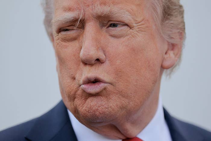 A person with a solemn expression is captured mid-speech in a suit and tie, against a plain background
