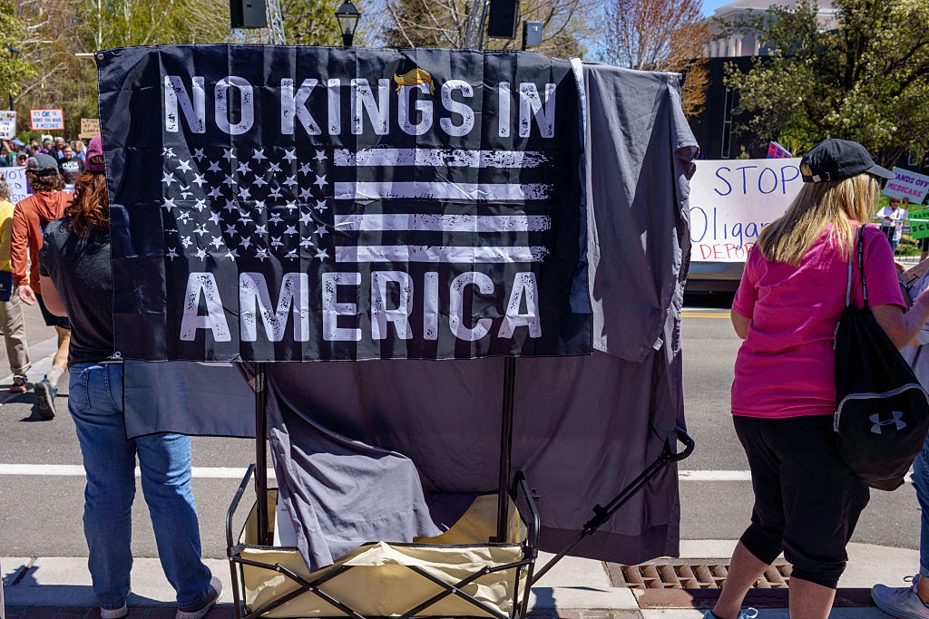 People at a protest with a sign reading "No Kings in America" featuring an altered U.S. flag. Other signs are partially visible in the background