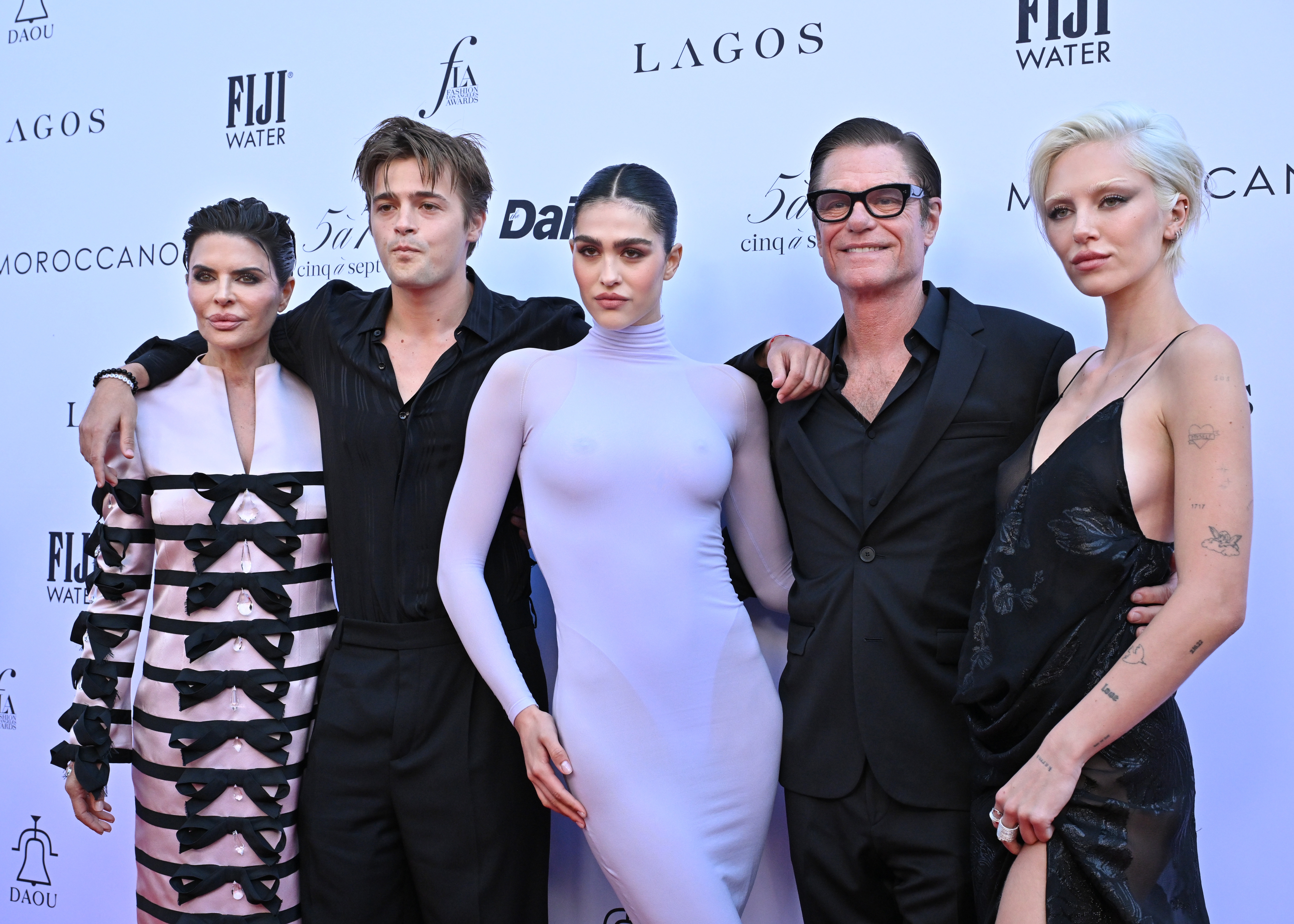 Five individuals pose on a red carpet, wearing stylish evening attire, including suits and elegant dresses