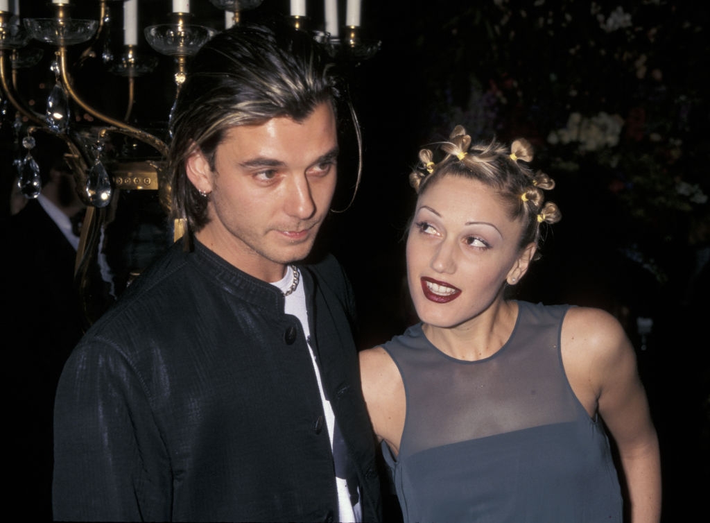 Two people at an event, one with a sleek jacket and another with a sleeveless top and twisty hairstyle, looking at each other