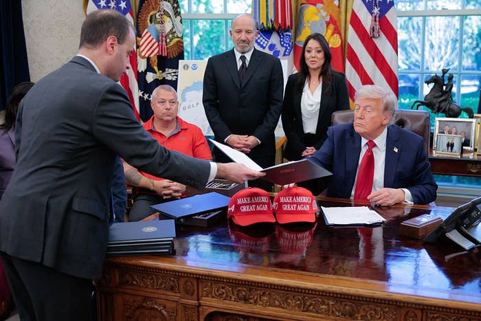 A man presents a document to a seated man in a suit behind a large desk, with several people standing nearby and "Make America Great Again" hats on the desk