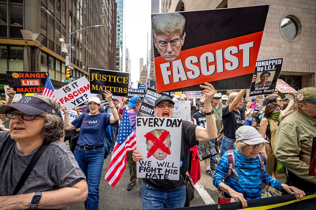 People protest in a city street, holding signs with messages like "Fascist" and "Every Day We Will Fight His Hate."