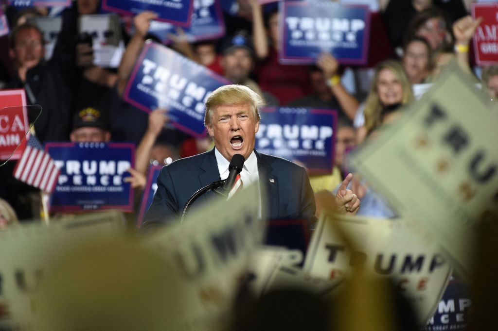 A person speaks passionately at a podium amid a cheering crowd holding signs at a political rally