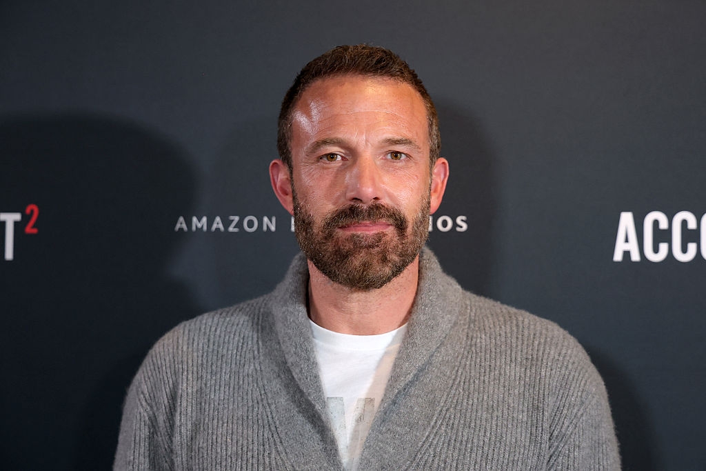 A person with short hair and a beard wears a casual sweater over a T-shirt at a media event