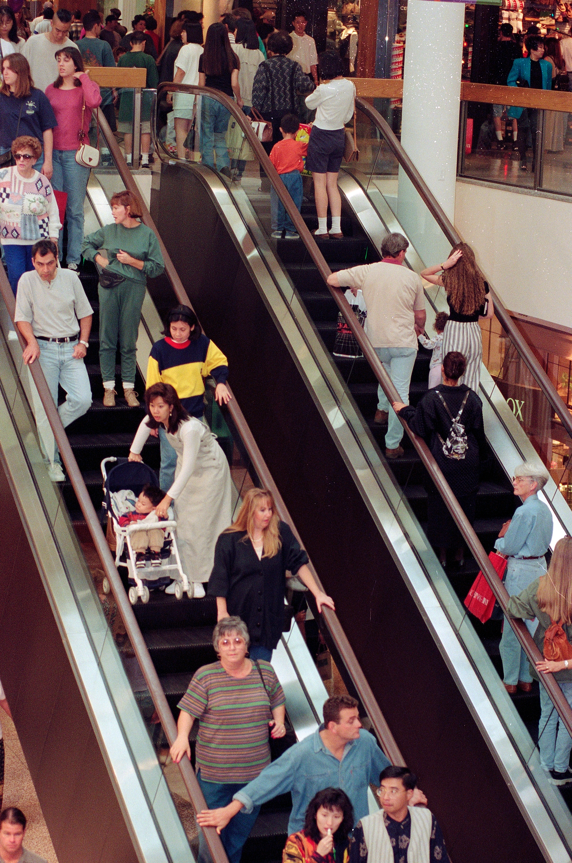 People on busy escalators in a shopping mall, some carrying bags and others pushing strollers