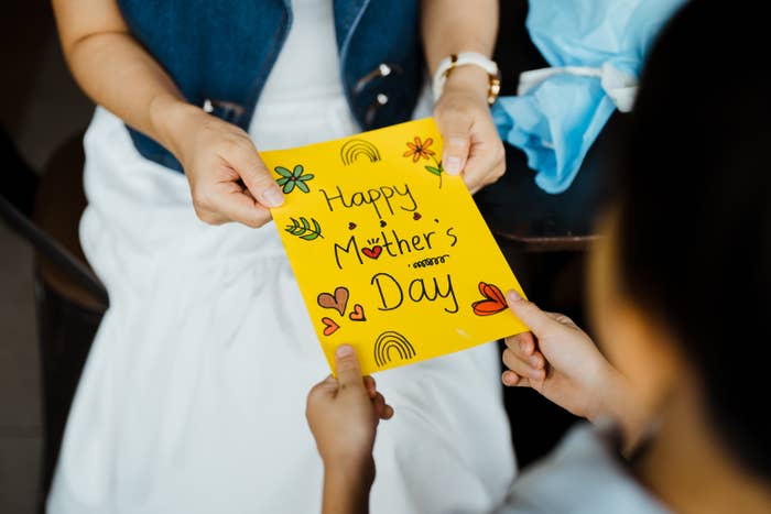 Child hands a colorful handmade “Happy Mother’s Day” card with flowers and hearts to an adult seated wearing a denim vest