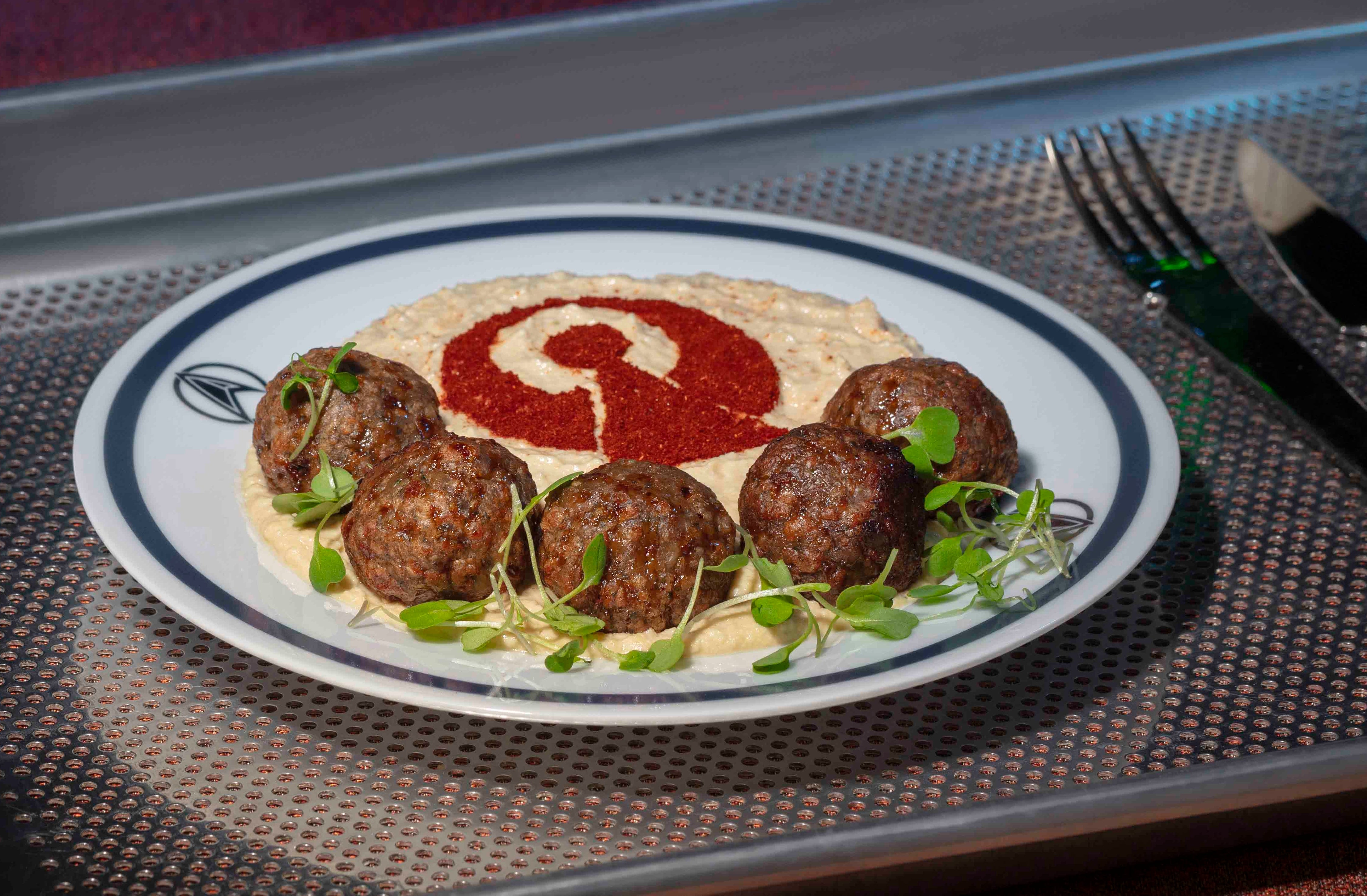 A plate with fritters and hummus, garnished with greens and a red swirl, is placed on a tray with a fork and knife beside it