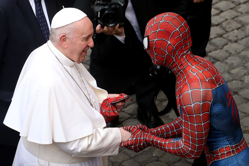 A person dressed as Spider-Man shakes hands with Pope Francis in white robes, surrounded by photographers