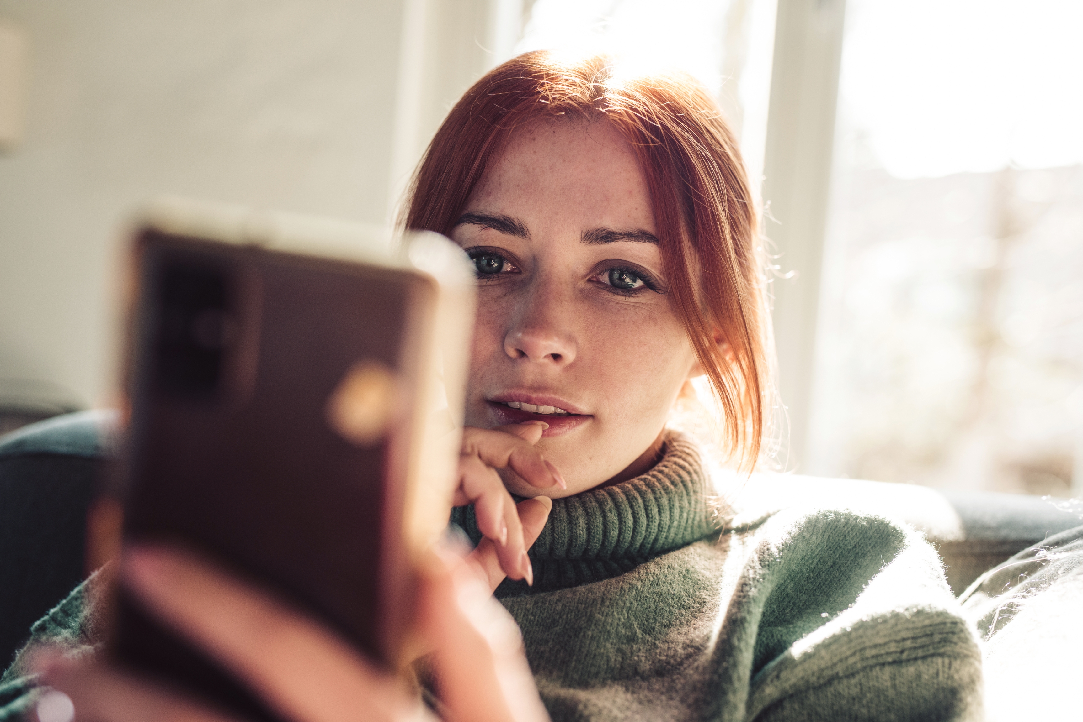 Person sitting, holding a smartphone, looking at the screen with a thoughtful expression, wearing a cozy sweater