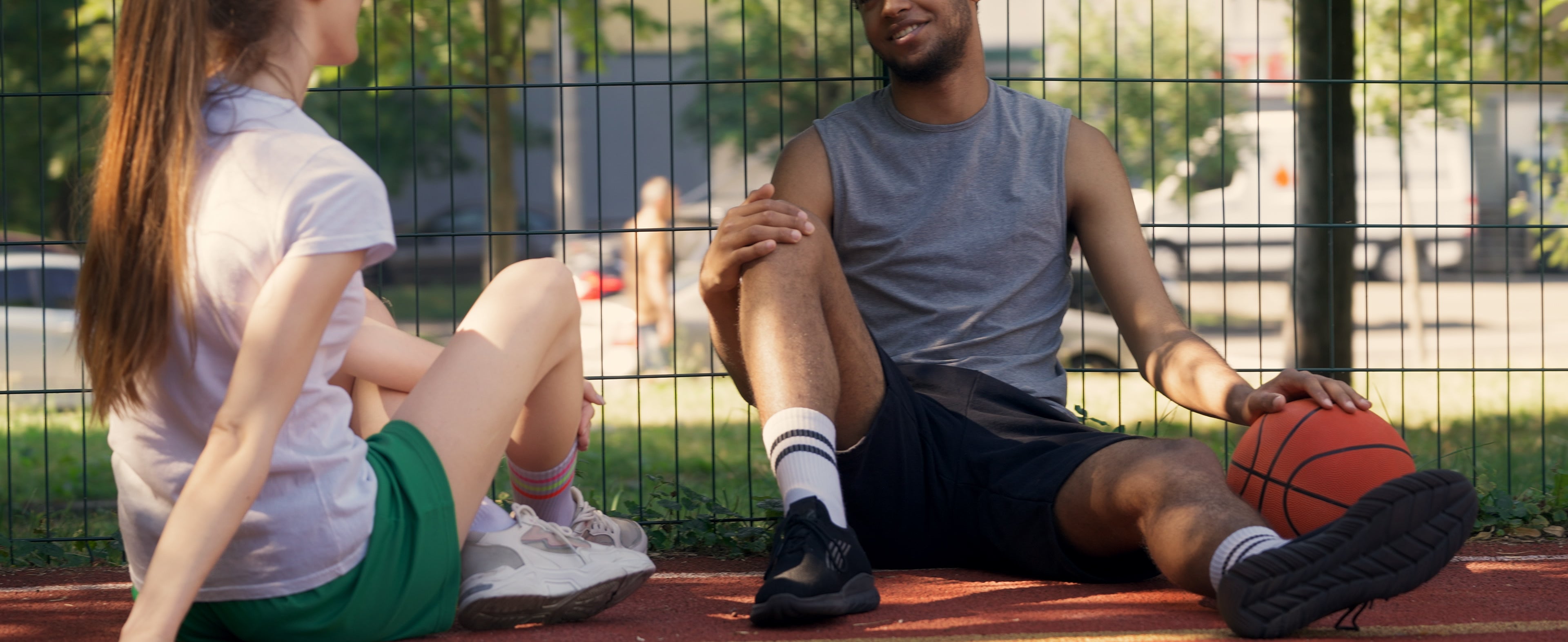 Two people, in casual athletic wear, chat and relax on a basketball court. One holds a basketball