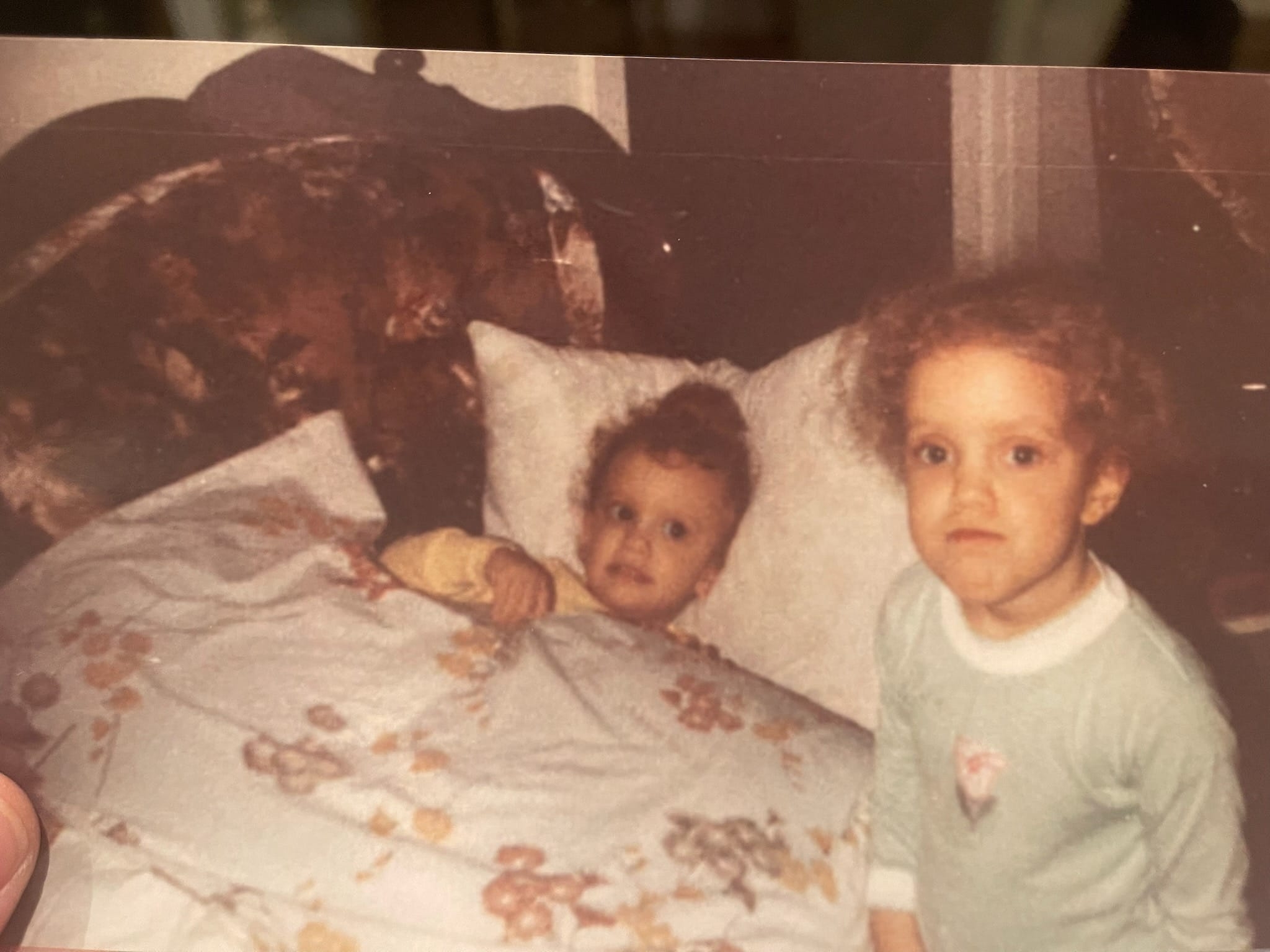 Two young children in a vintage bedroom setting; one is tucked in bed, while the other stands beside, both with curious expressions