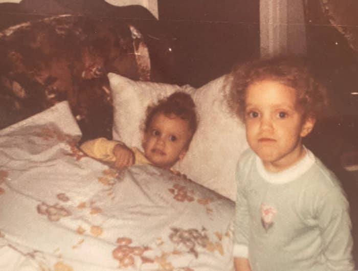 Two young children in a vintage bedroom setting; one is tucked in bed, while the other stands beside, both with curious expressions
