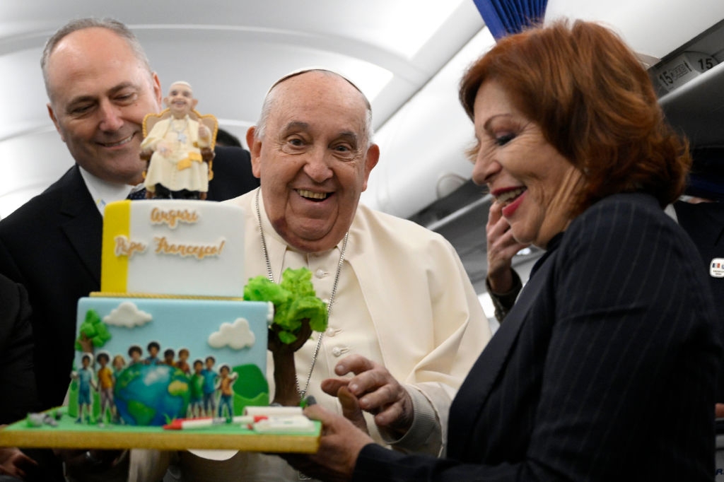 Pope Francis smiles while holding a cake featuring a globe, children, and a figure dressed similarly to him. Another person looks on happily