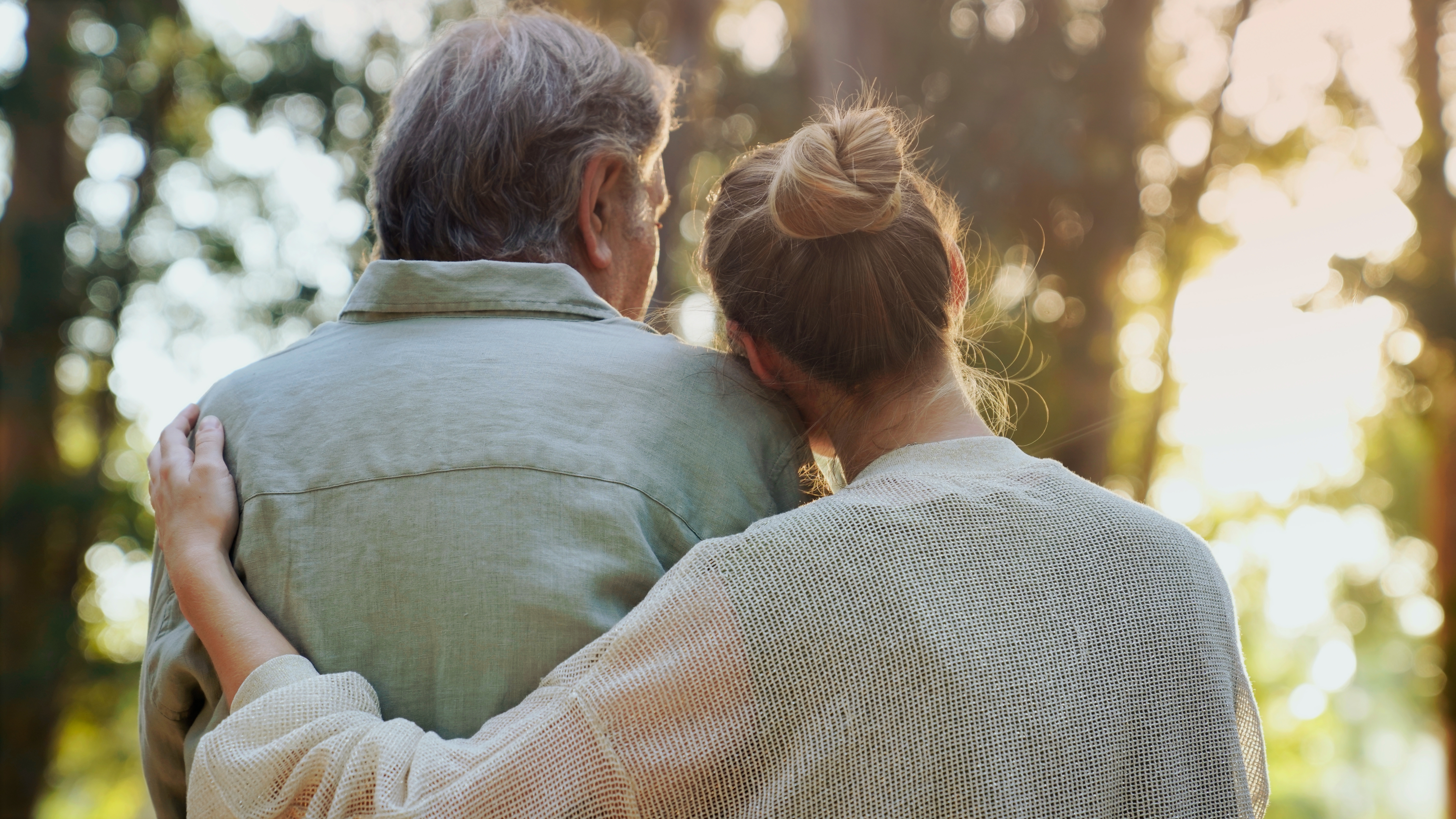 Older man and younger woman embracing while standing outdoors, woman resting her head on his shoulder