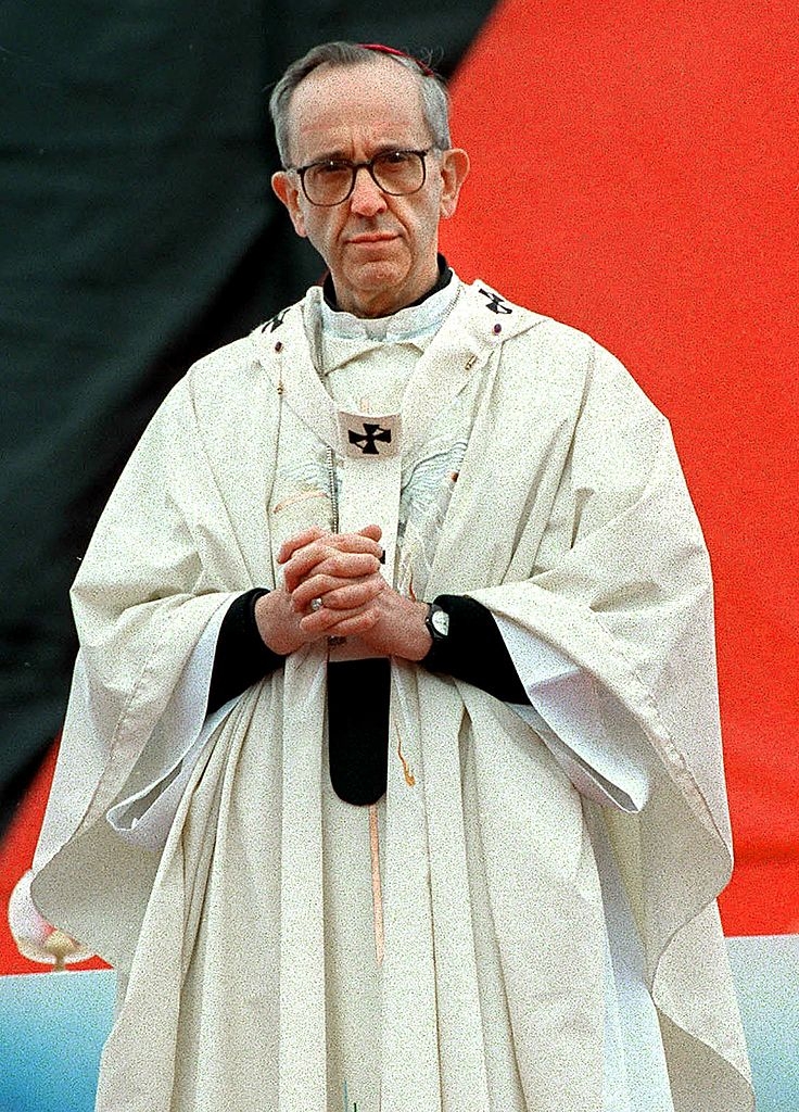 Archbishop Bergoglio in religious robes stands with hands clasped, wearing glasses and a serious expression