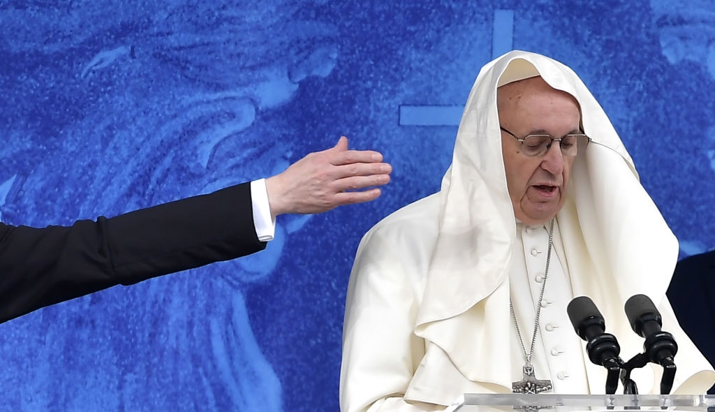 Pope Francis speaks at a podium with microphones, wearing traditional white robes and what appears to be a headpiece, but is a small cape draped over his head. A person's hand reaches out nearby to grab it