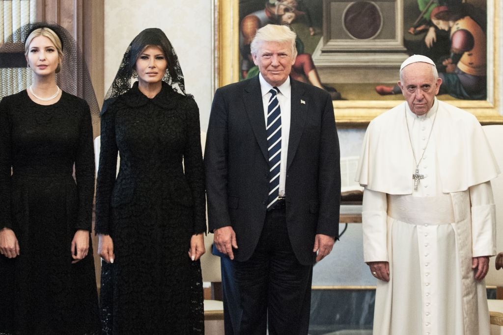 Pope Francis in religious garb, frowning, next to a smiling President Donald Trump; beyond President Trump are Melania and Ivanka Trump, both in black dresses with black veils