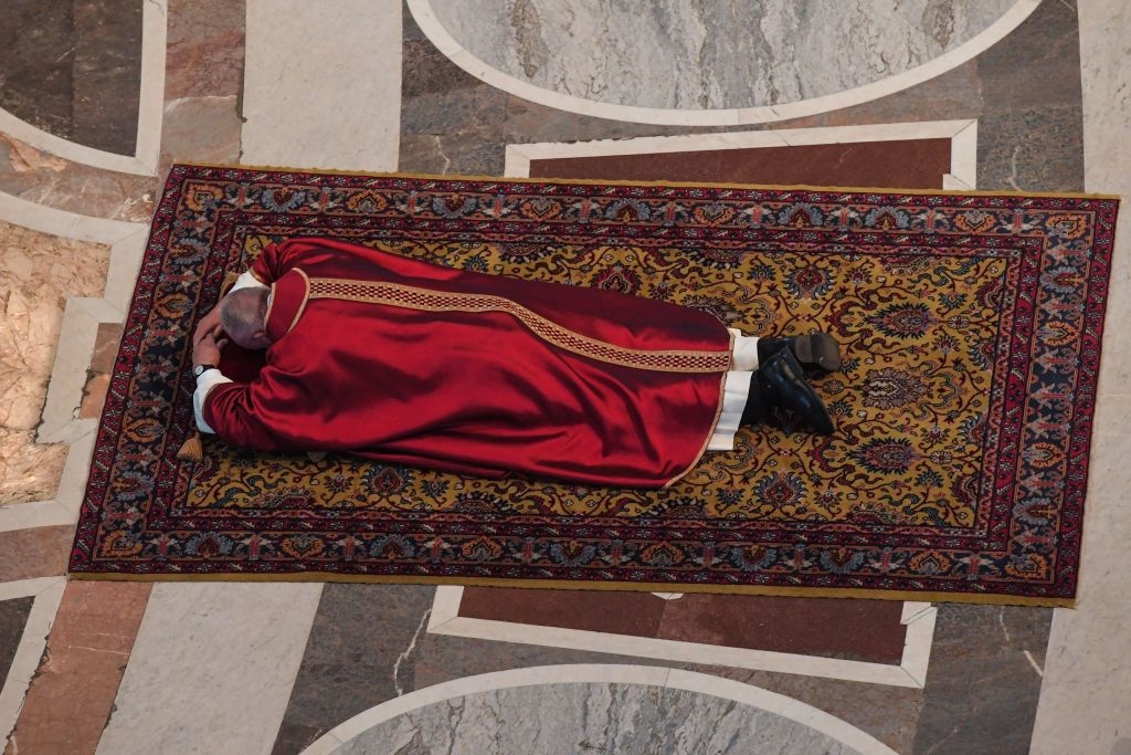 Pope Francis in religious robe lies prostrate on an ornate carpet during a solemn ceremony in a large hall