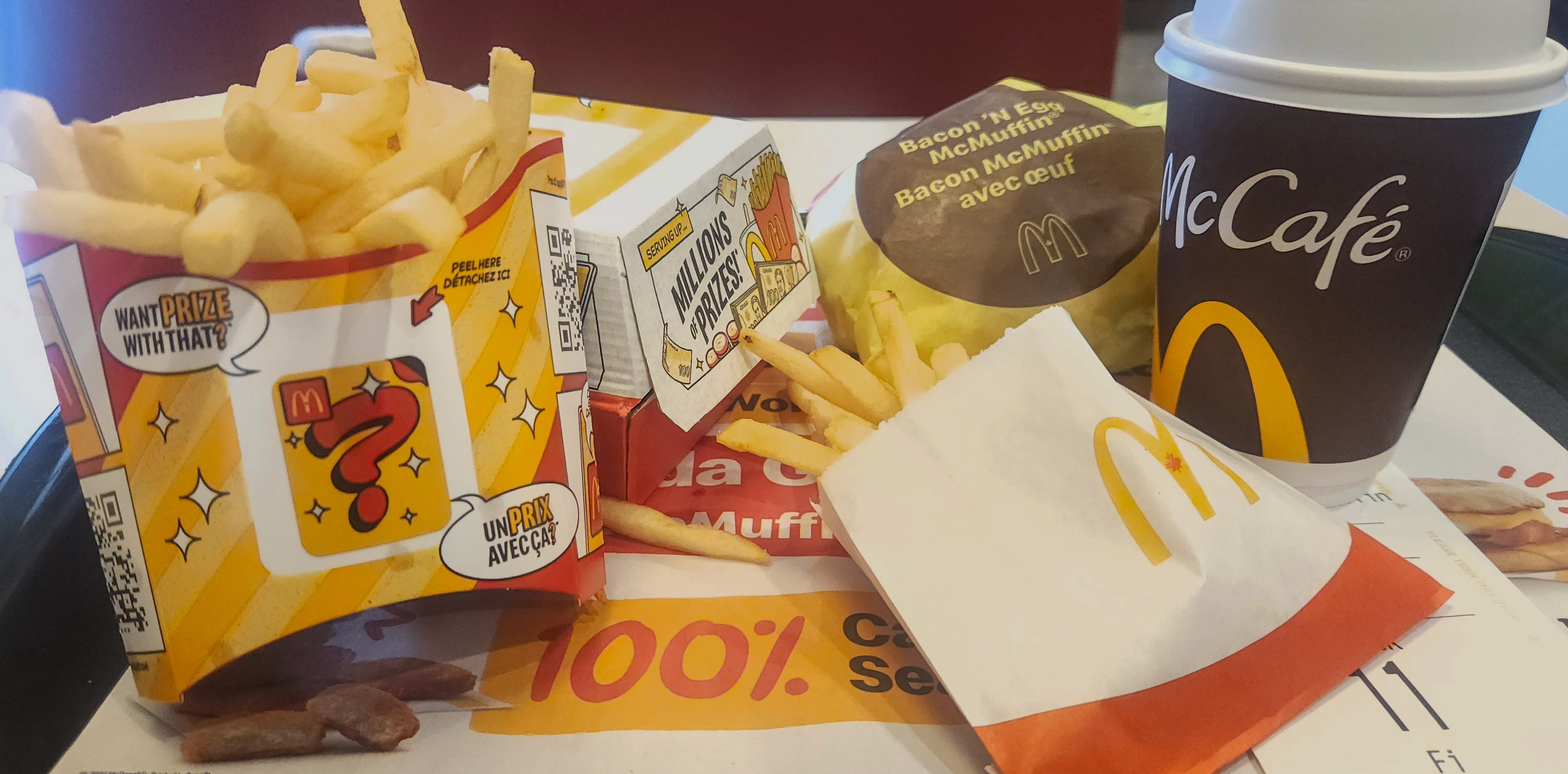Meal on a tray includes fries, two McDonald's Happy Meal boxes, a McMuffin, a McCafé coffee, and promotional text on the mat below