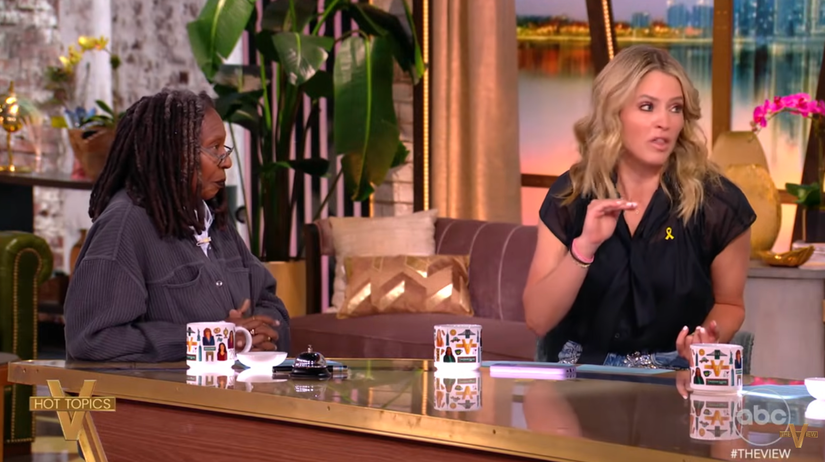 Two people are seated at a talk show panel discussing a topic. One person gestures while speaking. Coffee mugs with patterns are on the table