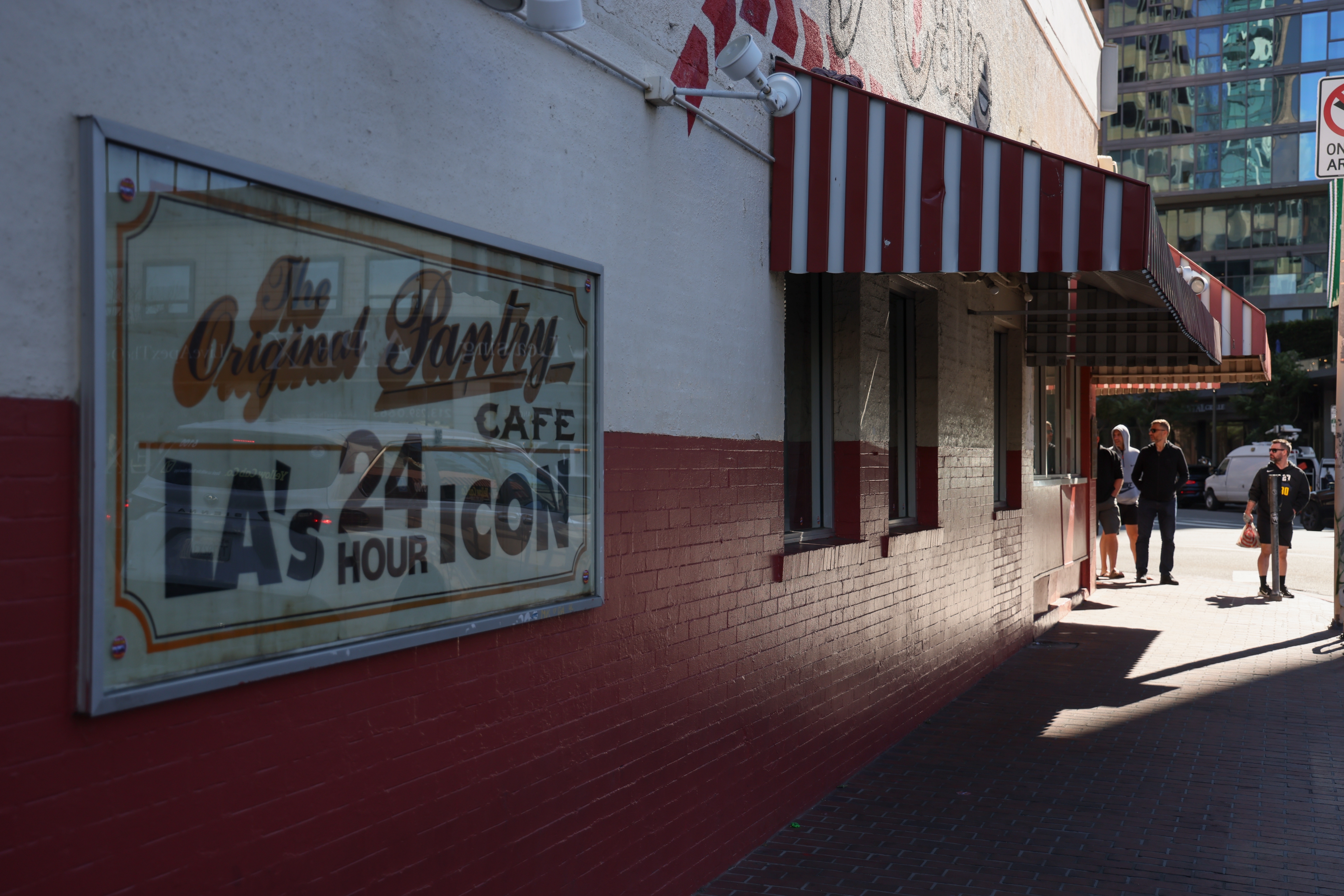 Sign for The Original Pantry Cafe, a 24-hour diner, on a building with a striped awning. Two people walk on the sidewalk nearby
