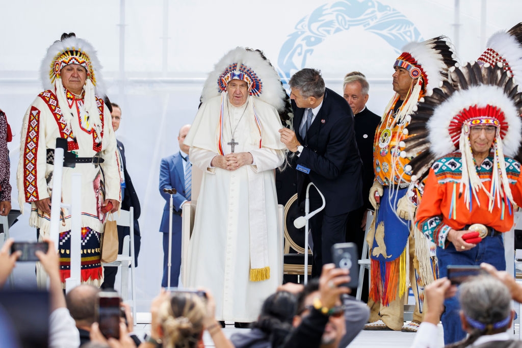 Pope Francis wearing a headdress stands among Indigenous leaders in traditional attire on a public stage