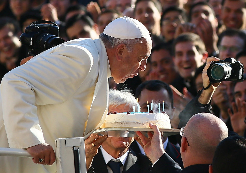Pope Francis leans over a birthday cake with lit candles, surrounded by a joyful crowd taking photos