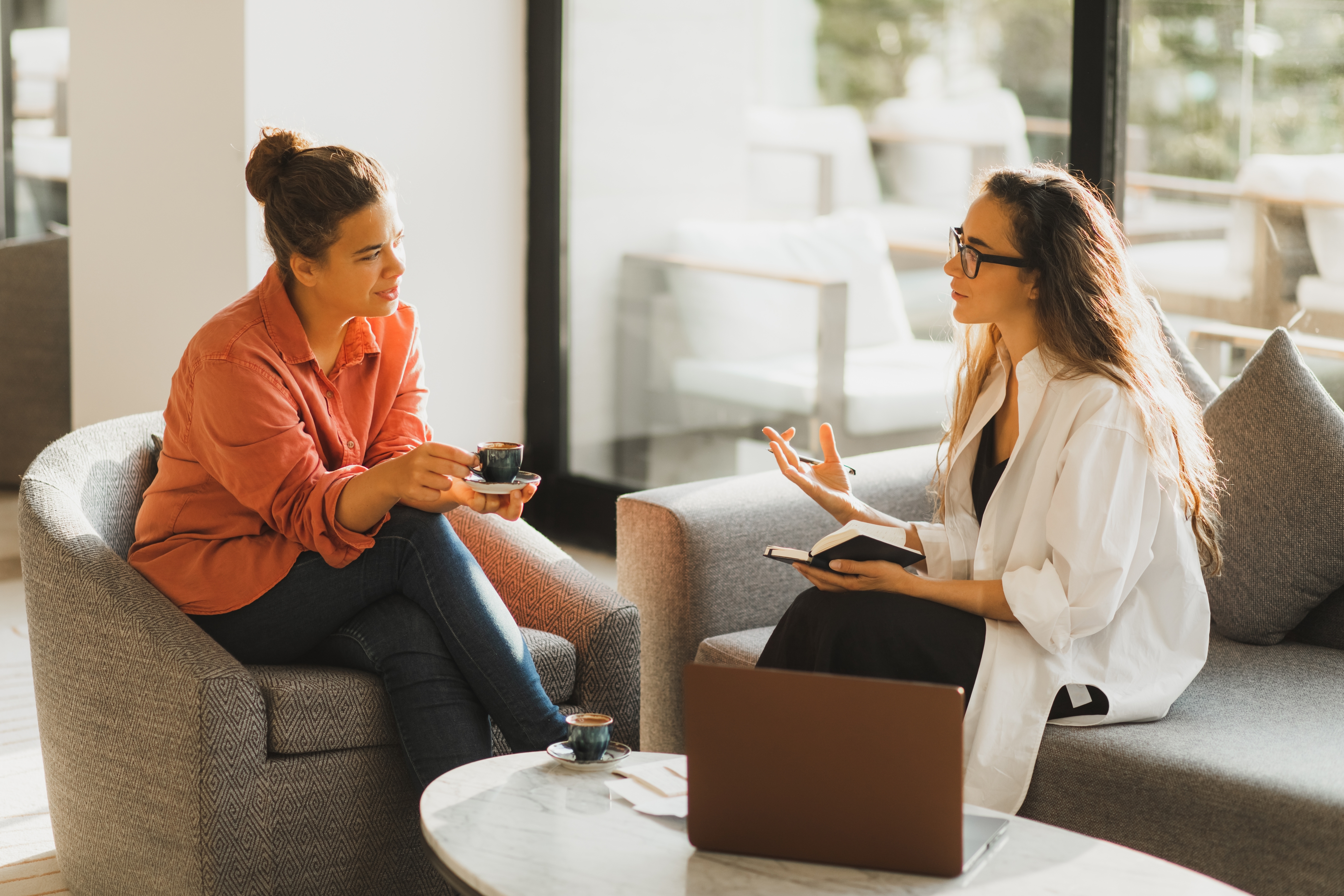 Two people are sitting on a couch, having a conversation indoors. One holds a cup and the other holds a notebook. A laptop is on the table