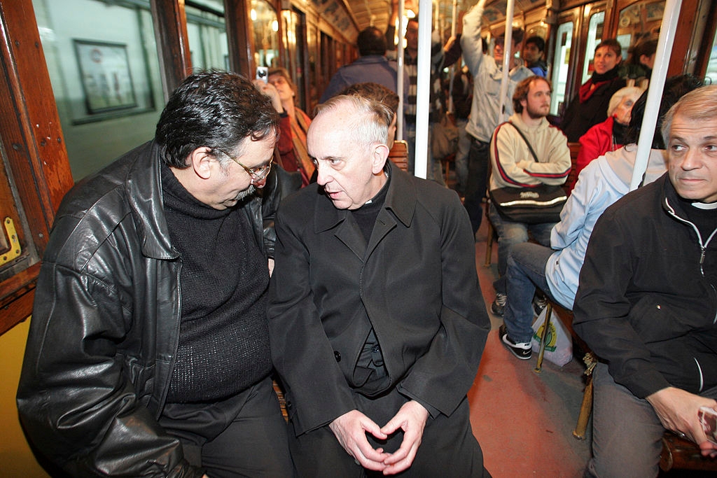 Cardinal Jorge Bergoglio in conversation on a crowded vintage subway car, surrounded by seated and standing passengers