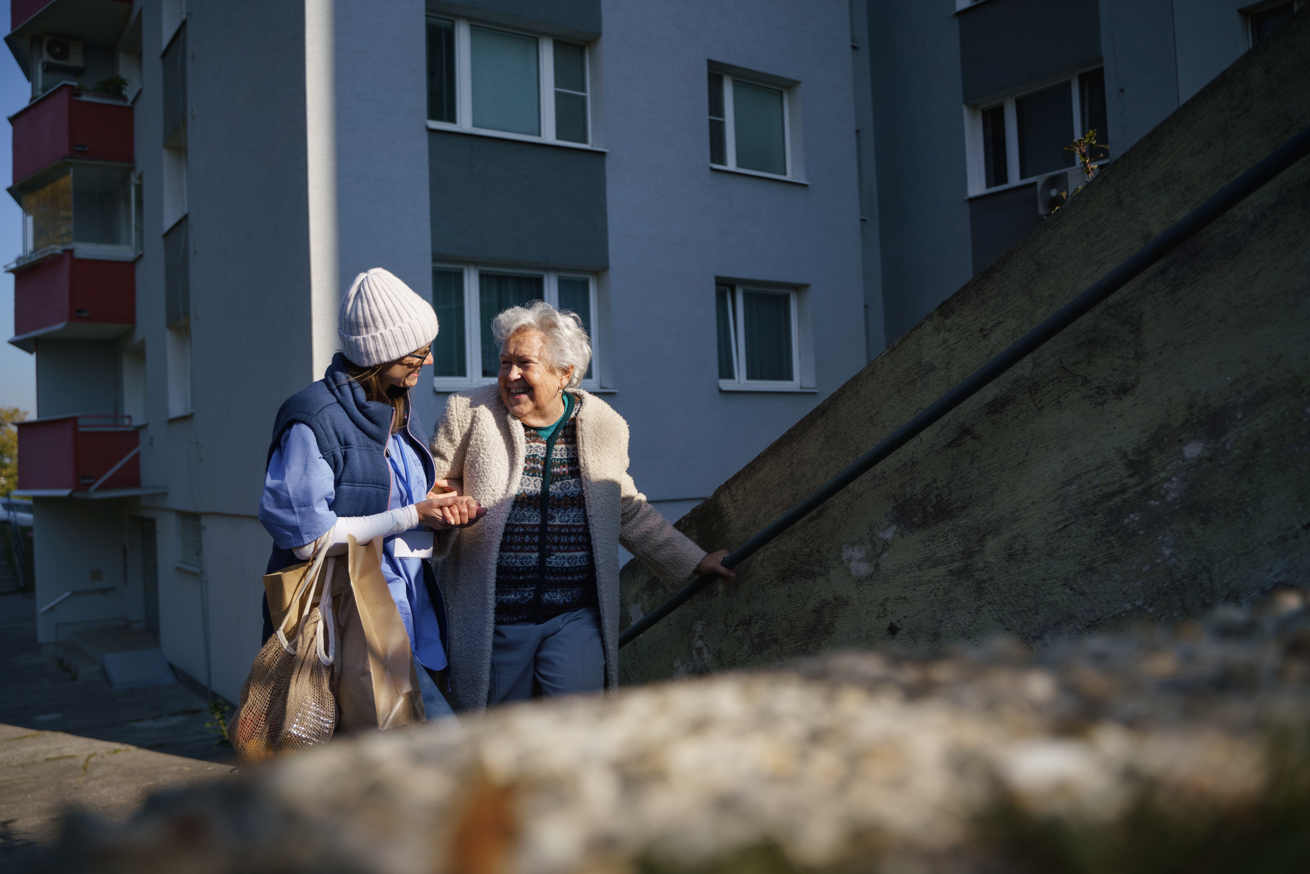 Two people walking outdoors near a building; one supports the other with a gentle touch and smile
