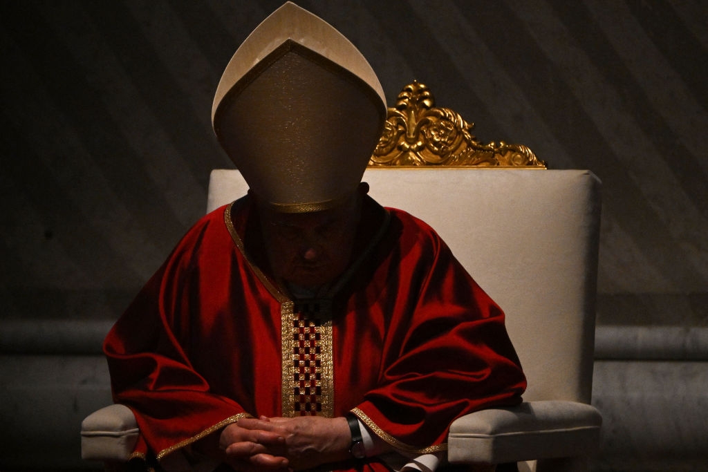 Pope Francis in ceremonial religious attire, including a large headdress, sits solemnly on a throne-like chair, with head bowed and hands clasped; his face is in shadow