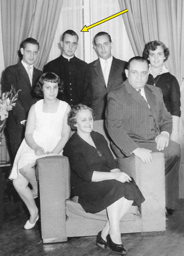 A group of seven people poses indoors, some seated and others standing, dressed in formal 1950s attire, with curtains in the background; an arrow points out Pope Francis standing in the back row