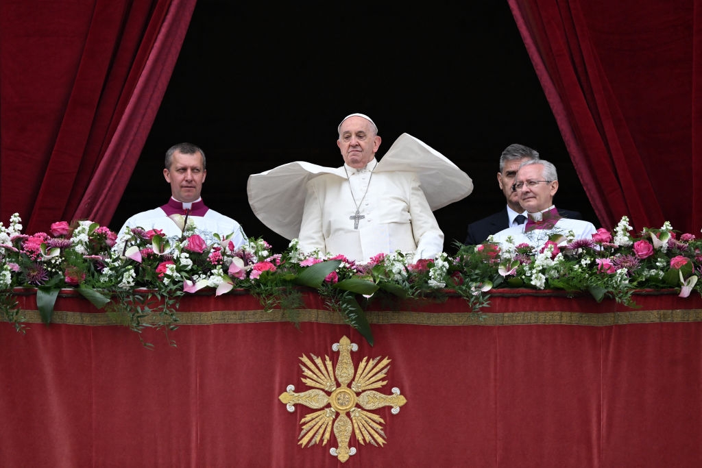 Pope Francis stands on a decorated balcony with three other clergy, surrounded by floral arrangements