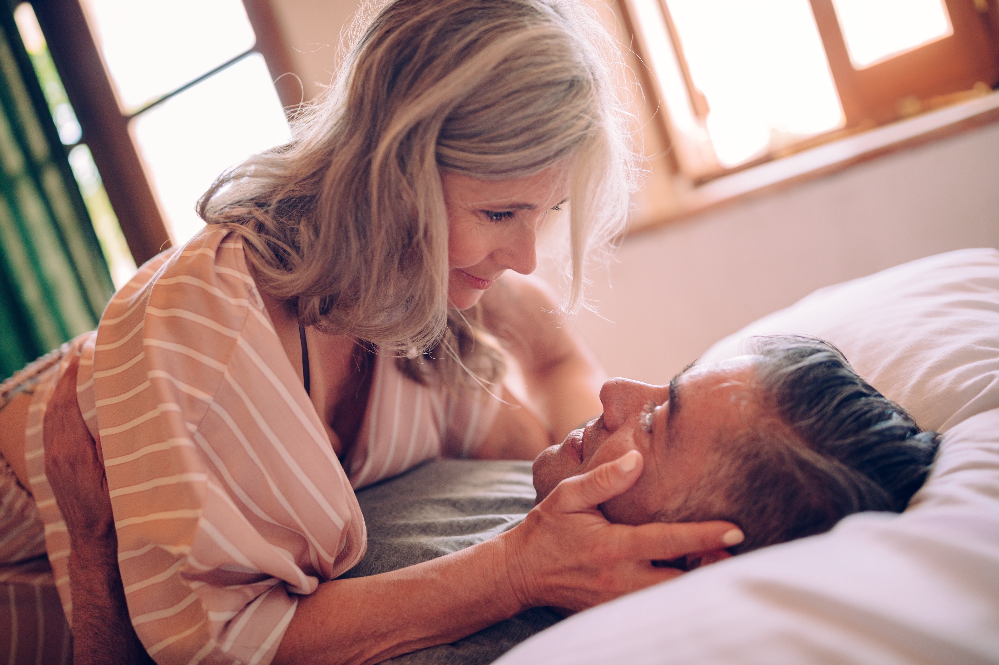 Older couple lovingly gazing at each other in bed, the woman propped above the man, smiling softly. The scene conveys intimacy and affection