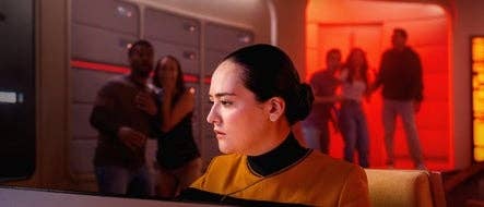 Person in futuristic uniform at control panel, focusing intently. In the background, people stand in a dimly lit room with glowing walls