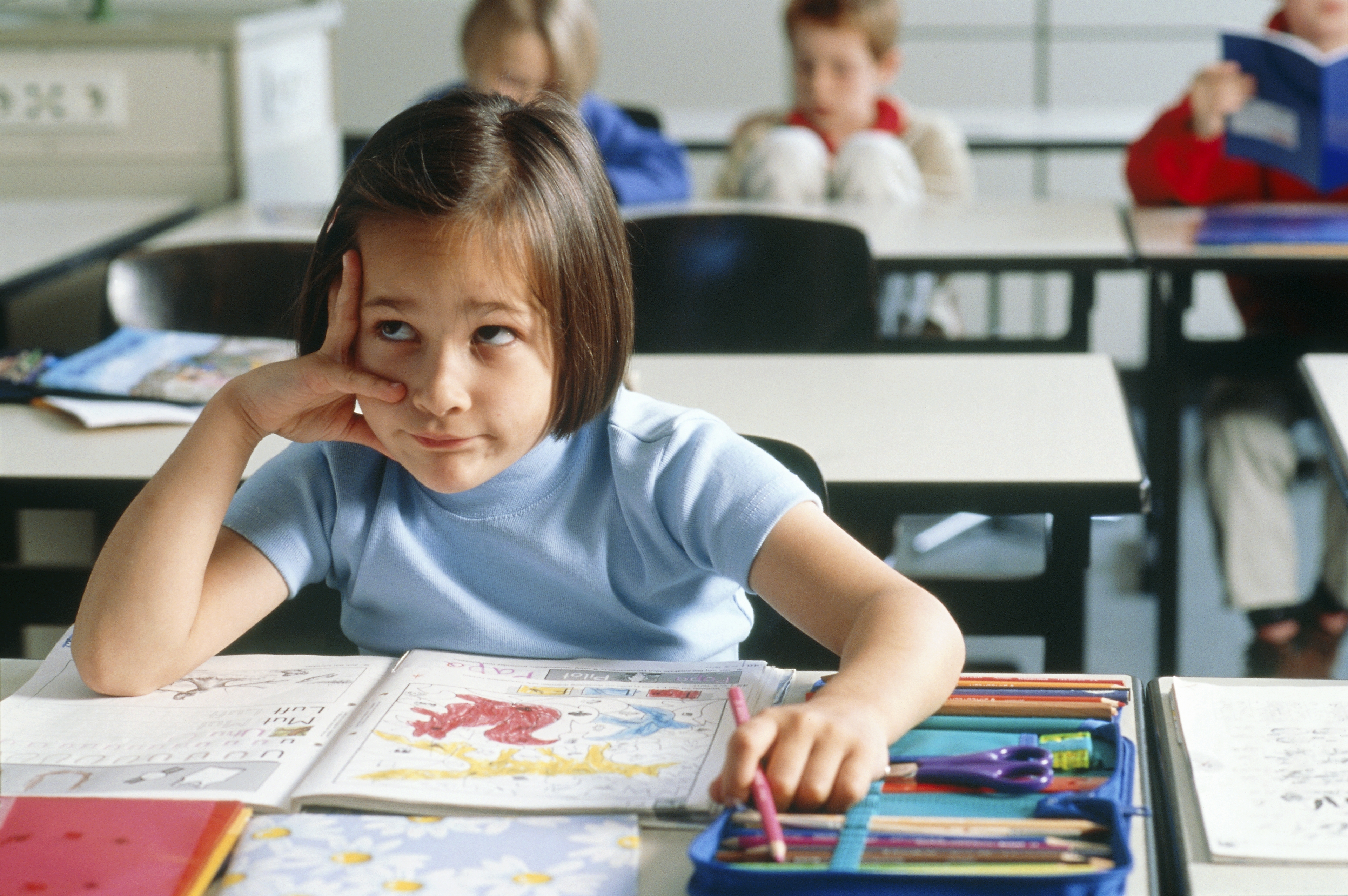 Child in a classroom looking bored while sitting at a desk with an open book and colored pencils. Other children are blurred in the background