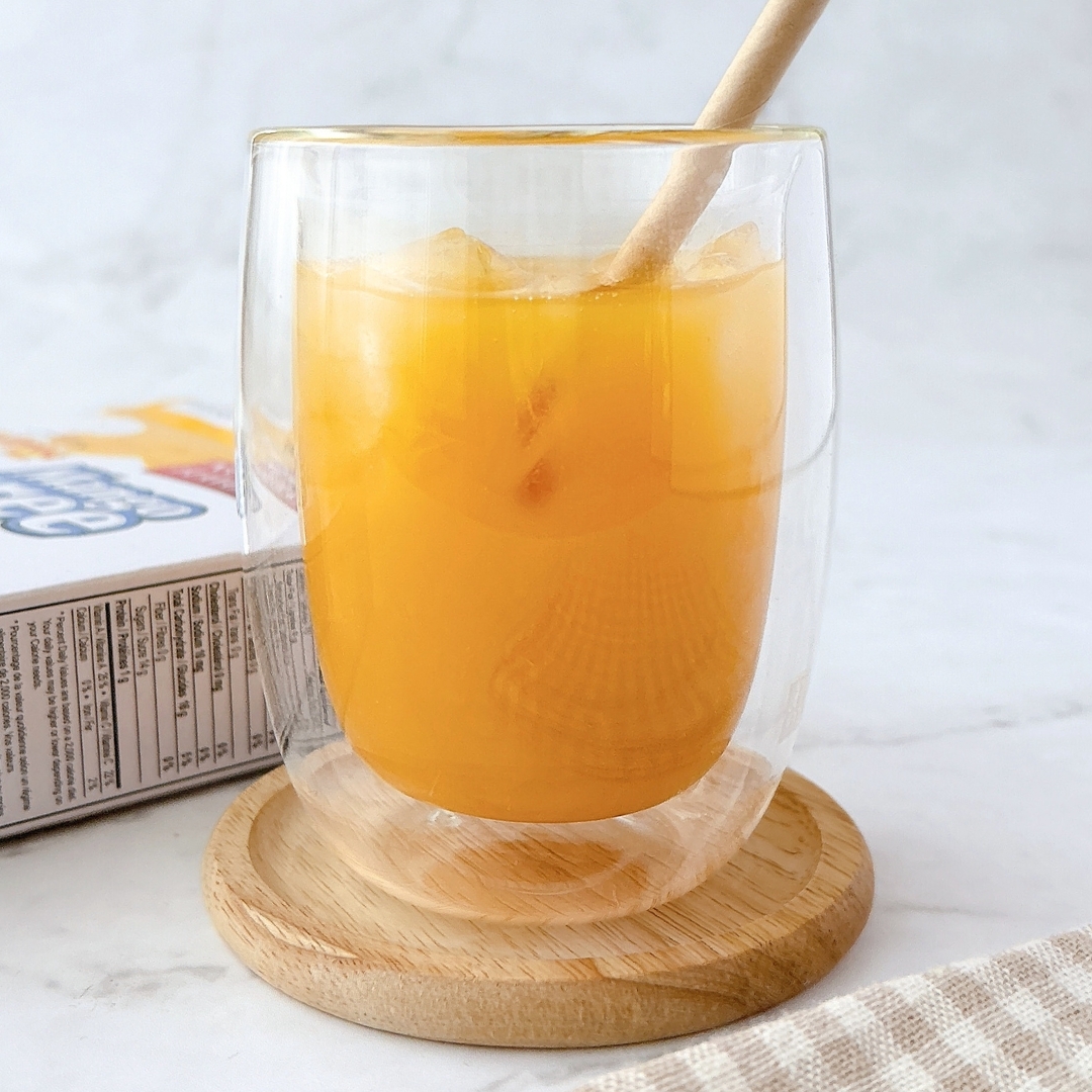 二重壁ガラスに入ったオレンジジュース。木製のコースターに置かれ、ストローが刺してある。背景にジュースの箱が見える。