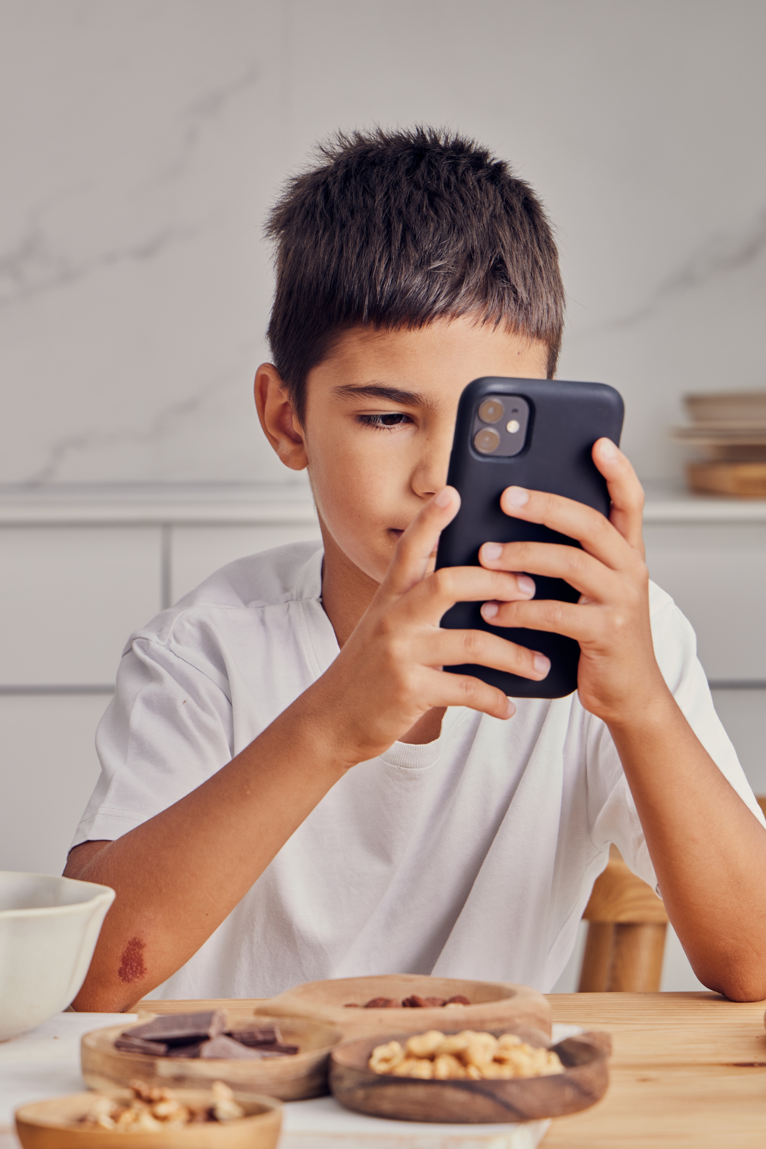 Child in a white shirt intently using a smartphone at a kitchen table with bowls of food in front of him